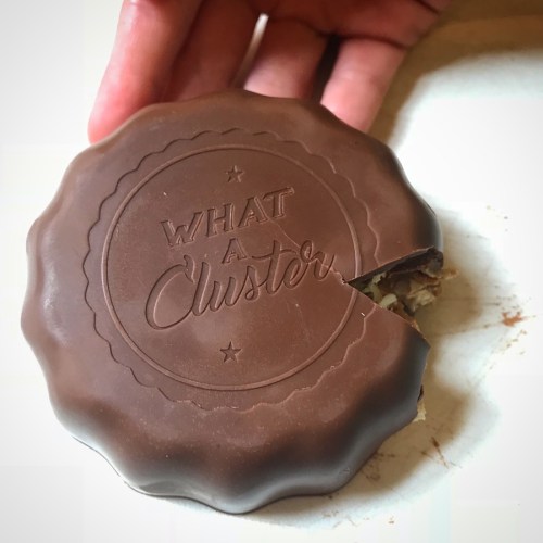 A hand holds a chocolate-covered treat with "WHAT A CLUSTER" embossed on top, showing a bite taken out.