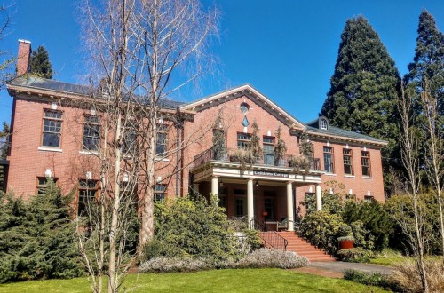 Historic brick building with a porch, surrounded by greenery and tall trees under a clear blue sky.