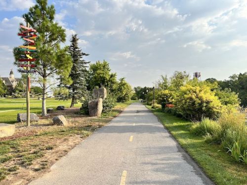 A paved path lined with greenery and rocks, leading through a park with colorful sculptures and trees under a cloudy sky.