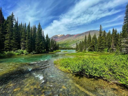 A serene river flows through a lush green landscape, surrounded by tall trees and mountains under a blue sky.
