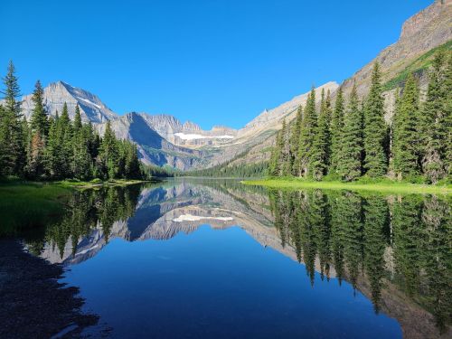 A serene mountain landscape reflected in a calm lake, surrounded by lush green trees under a clear blue sky.