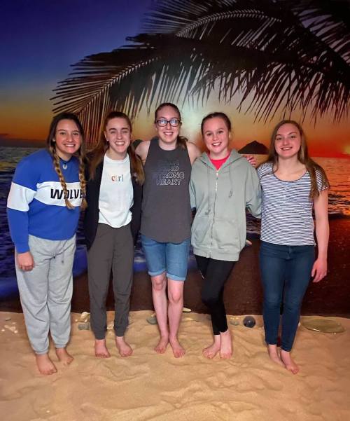 Five young women stand barefoot on sand, smiling in front of a beach sunset backdrop with palm trees.