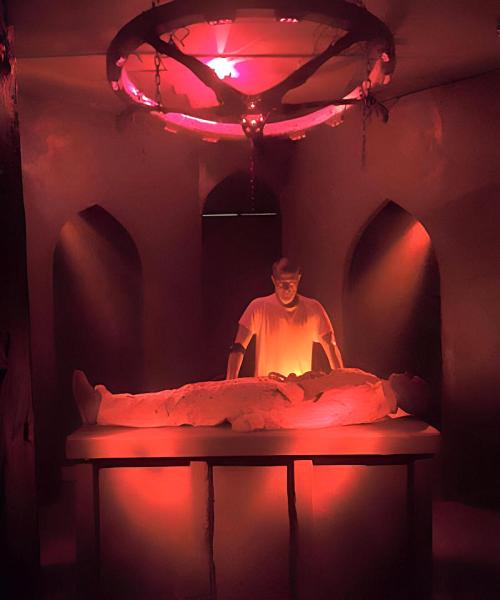 A dimly lit room with a figure standing over a person lying on a table, illuminated by red light and a circular chandelier.