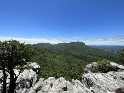A panoramic view of lush green mountains under a clear blue sky, with rocky outcrops in the foreground.