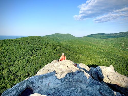 A person sits on a rocky ledge overlooking a lush green valley and distant mountains under a clear blue sky.