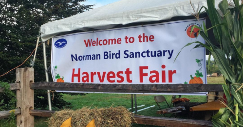 a white vinyl sign with blue and red lettering stating 'welcome to the norman bird sanctuary harvest fair" which occurs annually in middletown rhode island