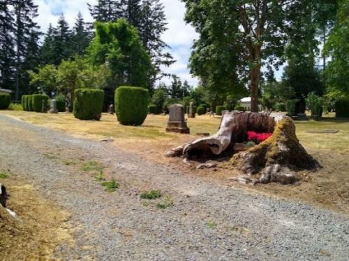 A gravel path in a cemetery lined with trees and neatly trimmed bushes, featuring a large tree stump and a flower arrangement.