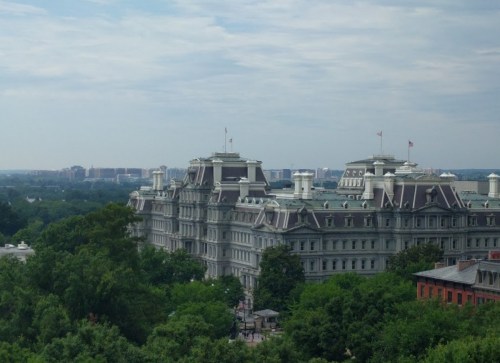 A historic building with a gray facade and multiple flags, surrounded by trees and a distant city skyline under a cloudy sky.