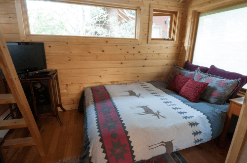 Cozy interior of a tiny cabin featuring a bed with decorative pillows, a TV, and wooden walls.