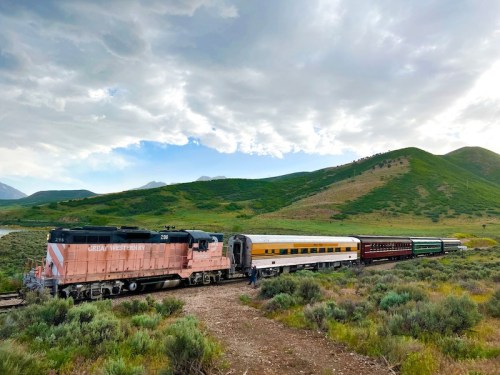 A vintage train with multiple cars travels through a lush green landscape under a cloudy sky.