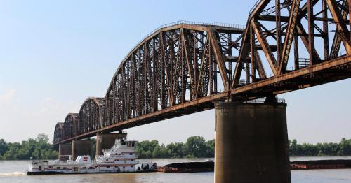 A rusty bridge spans a river, with a barge passing underneath, surrounded by green trees and a clear sky.