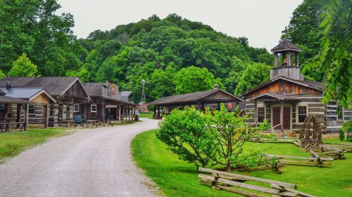 A scenic view of a rustic village with wooden buildings along a gravel path, surrounded by lush green trees.