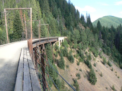 A scenic view of a curved bridge surrounded by lush green trees and mountains under a clear blue sky.