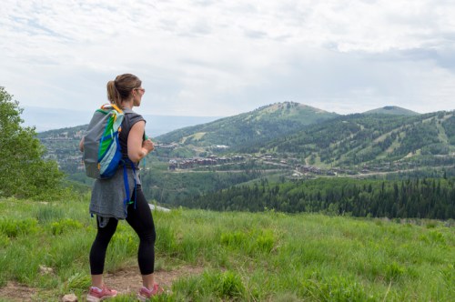 A woman stands on a grassy hill, looking out over a lush green valley and distant mountains.