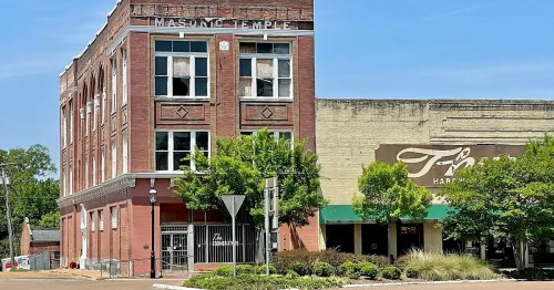 Historic brick building labeled "Masonic Temple" next to a hardware store, with trees and clear blue sky in the background.