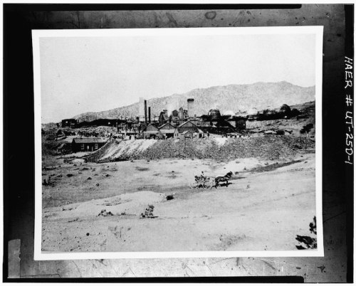 Black and white photo of an industrial site with buildings and smokestacks, set against a mountainous landscape.