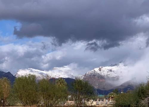 Snow-capped mountains under a cloudy sky, with greenery in the foreground and mist rising around the peaks.