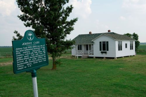 Historic marker for Johnny Cash in front of a white house, set in a grassy field under a cloudy sky.