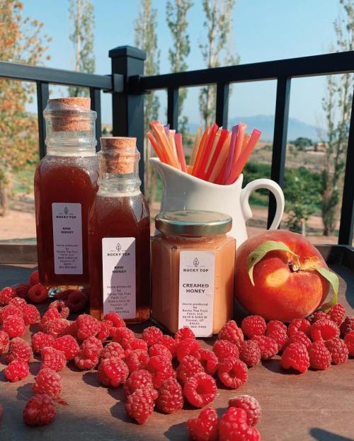 A wooden tray with jars of honey, creamed honey, fresh raspberries, a peach, and colorful straws against a scenic backdrop.