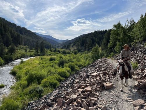 A person on horseback rides along a rocky path beside a river, surrounded by lush green hills and trees under a blue sky.