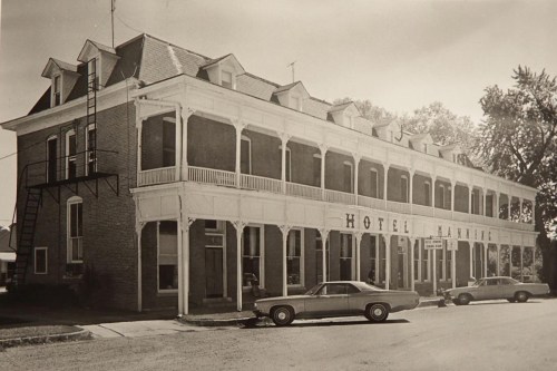 Historic hotel with a white wooden porch, vintage cars parked outside, and a fire escape on the side. Black and white photo.