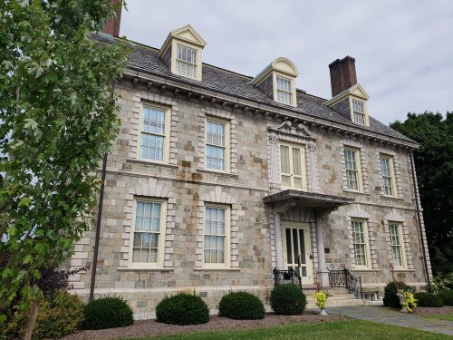 Historic stone house with a symmetrical facade, large windows, and a decorative entrance, surrounded by greenery.