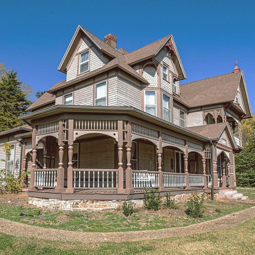 A large, historic Victorian-style house with a wraparound porch and intricate architectural details against a clear blue sky.