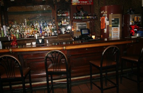 A dimly lit bar with a wooden counter, empty chairs, and shelves lined with various liquor bottles.