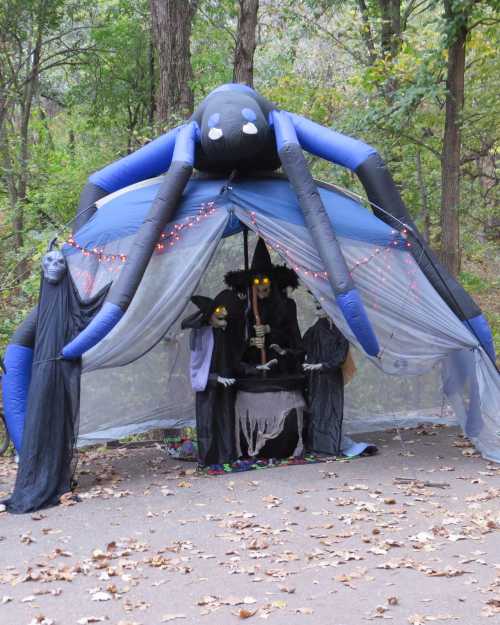 A large inflatable spider sits atop a spooky Halloween display with figures in costumes under a draped canopy.