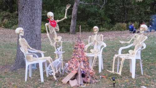 Four skeletons sitting around a campfire, decorated with lights, in a wooded area.