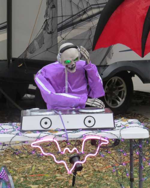 A skeleton DJ in a purple outfit spins records at a Halloween-themed outdoor setup with colorful lights.