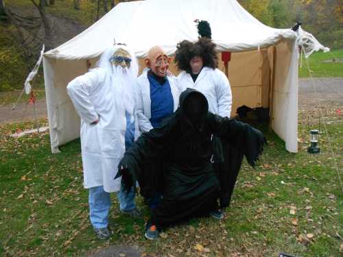 Four people in costumes, including a grim reaper and various masks, pose in front of a tent in a wooded area.