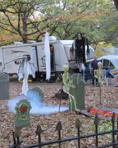 A spooky Halloween scene with skeletons and ghosts near a camper, surrounded by gravestones and autumn leaves.