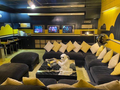 A stylish living room with a black sectional sofa, yellow accents, multiple TVs, and a decorative white alien figure.