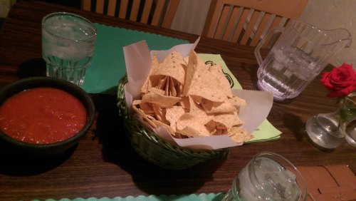 A basket of tortilla chips with a bowl of salsa, two glasses of water, and a rose on a table.