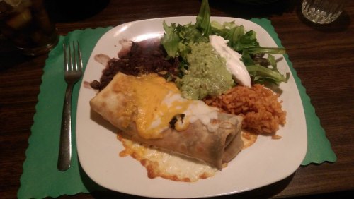 A plate of a burrito topped with cheese, served with rice, black beans, guacamole, sour cream, and a side salad.