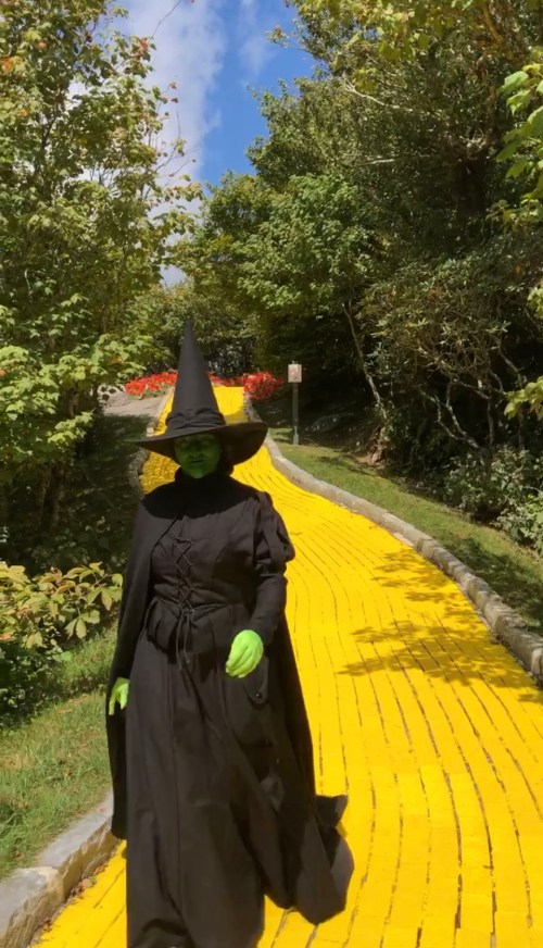 A person dressed as a witch with green skin walks along a bright yellow brick road surrounded by trees.