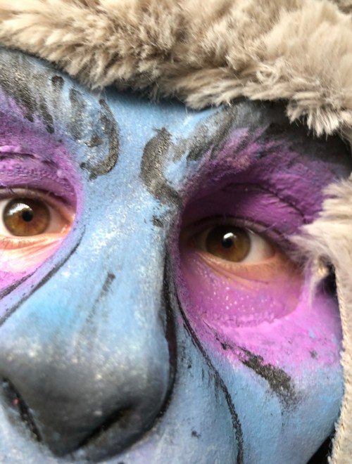 Close-up of a face painted in blue and purple, with expressive eyes and a furry hat.