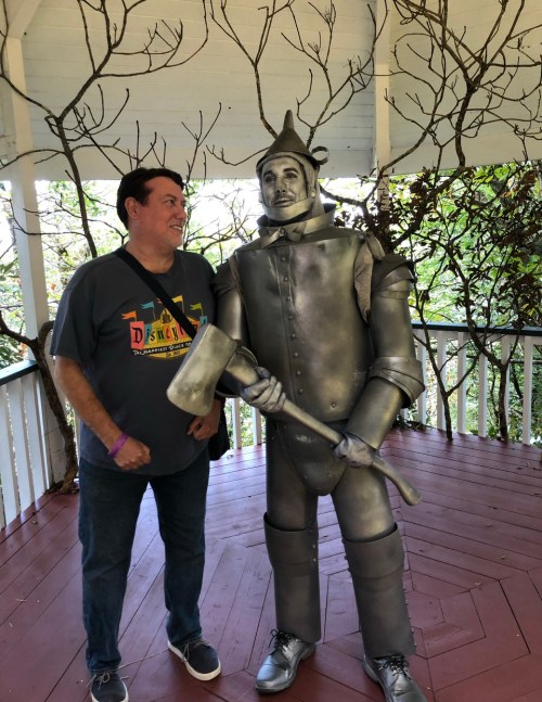 A man stands next to a silver Tin Man statue, both smiling, in a gazebo surrounded by trees.