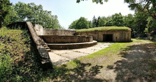Abandoned concrete structure with grassy roof, surrounded by trees and a clear blue sky. Steps lead to a circular platform.