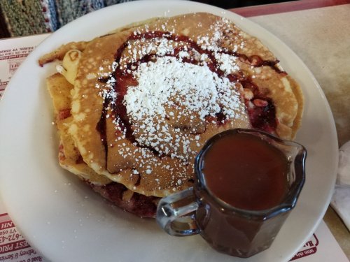 A stack of pancakes topped with powdered sugar and fruit filling, served with a small pitcher of syrup.