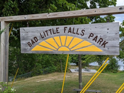 Sign for Bad Little Falls Park featuring a yellow sun design against a wooden background, surrounded by greenery.