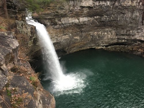 A waterfall cascades into a deep, green pool surrounded by rocky cliffs and sparse vegetation.