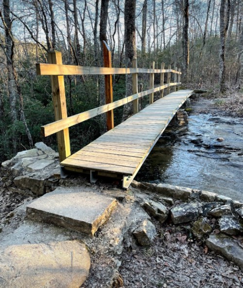A wooden bridge spans a small stream, surrounded by trees and rocky terrain in a serene outdoor setting.
