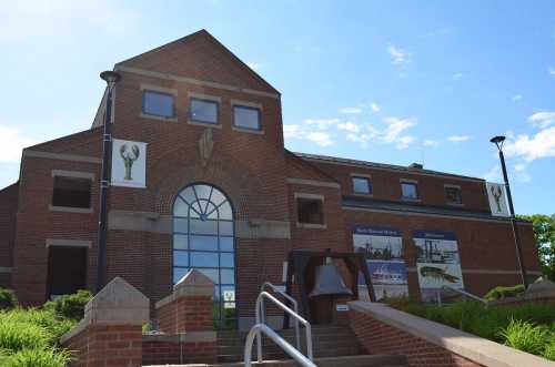 A brick building with large windows, featuring banners and a bell in front, under a clear blue sky.