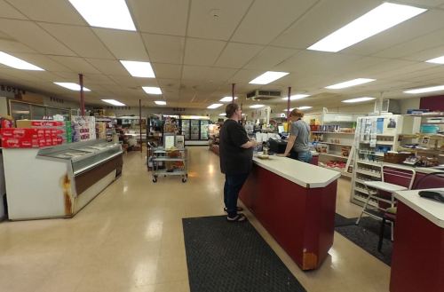 A grocery store interior with a checkout counter, shelves of products, and two customers interacting.
