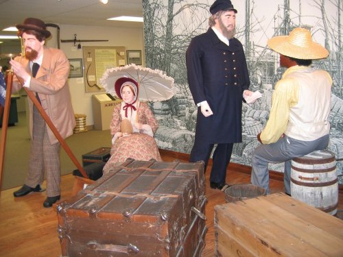 Historical scene with life-sized figures in period clothing, including a woman with a parasol and men in hats, near wooden crates.