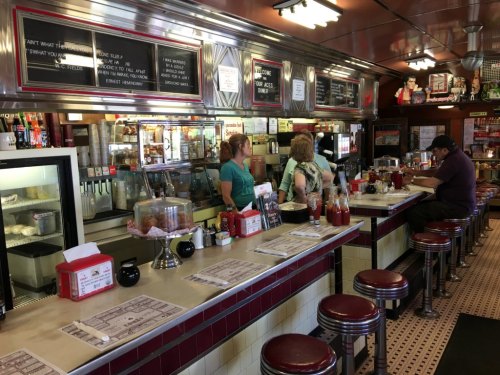 A retro diner interior with a long counter, patrons seated, and a menu board above. Classic decor and food items visible.