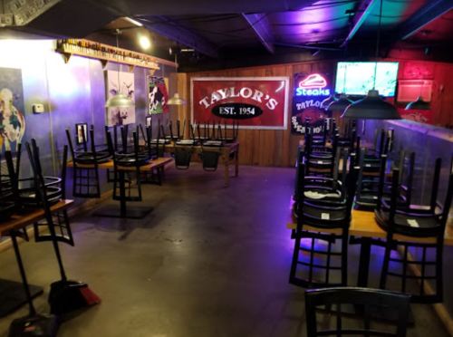 Empty restaurant interior with stacked chairs on tables, dim lighting, and a sign for "Taylor's" on the wall.