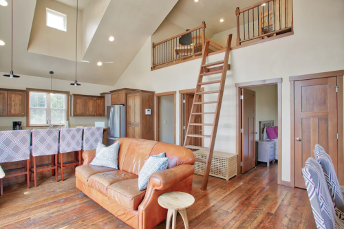 Cozy living area with a leather sofa, wooden accents, and a lofted space accessible by a staircase. Bright and inviting.
